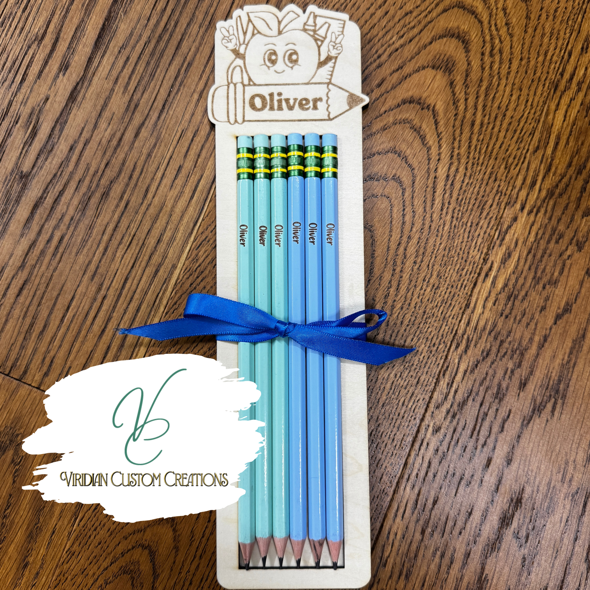 A set of five personalized light blue pencils in a wooden pencil holder, tied with a blue ribbon, displayed on a wooden table. The top of the holder is customized with a cartoon Apple and the name 'Oliver'.