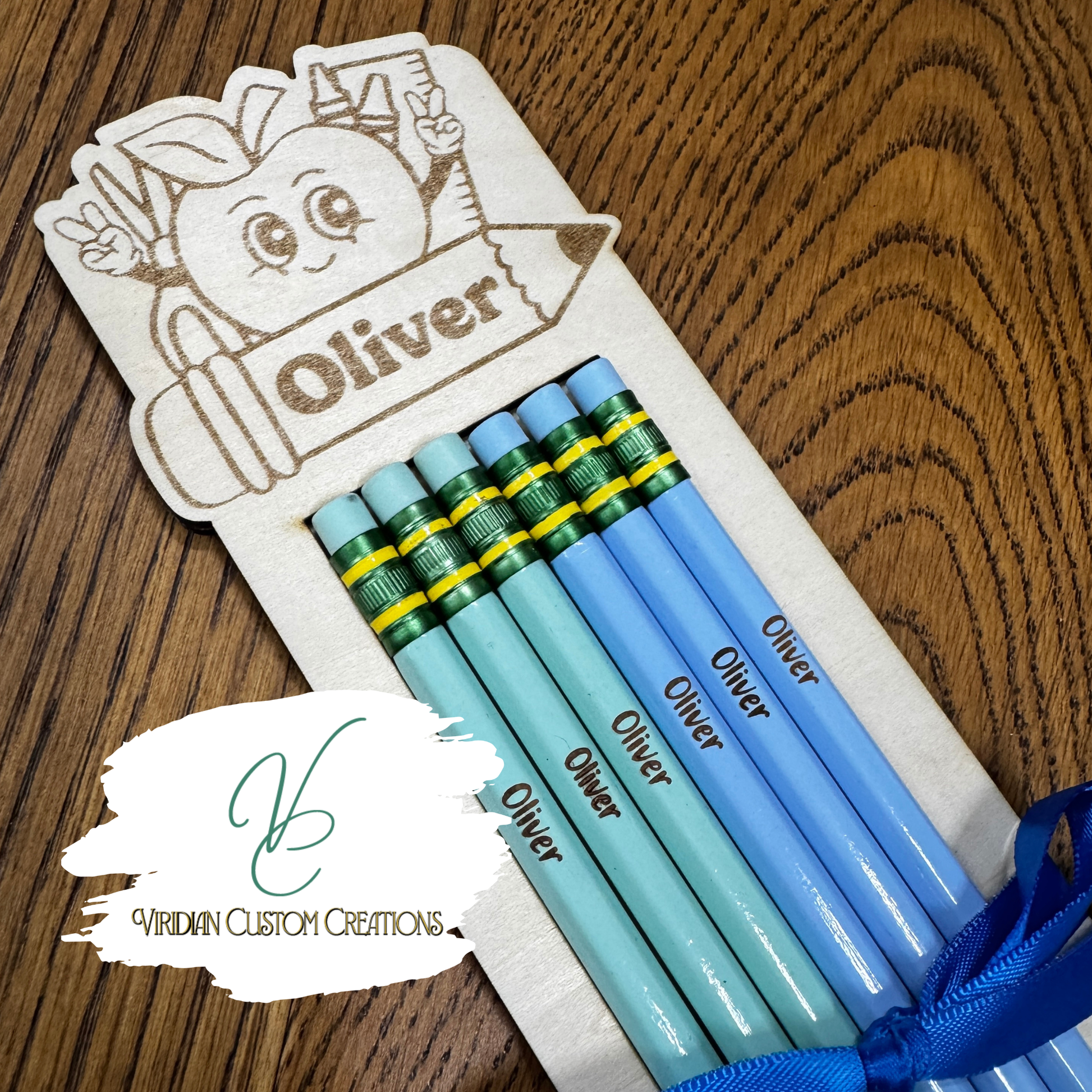 Five personalized blue pencils with green and yellow ferrules, custom engraved with the name 'Oliver', presented in a wooden pencil holder on a wooden table. A watermark for 'Viridian Custom Creations' is visible in the lower-left corner.