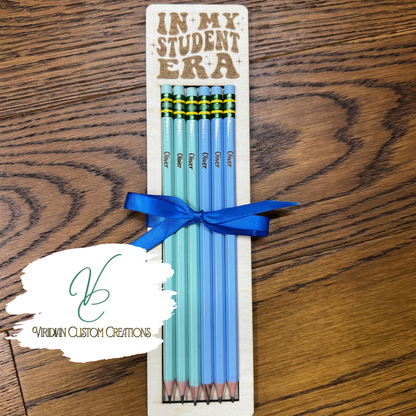 A set of six custom 'In My Student Era' pencils tied with a blue ribbon, presented on a wooden card, placed on a wooden tabletop. A watermark logo for 'Viridian Custom Creations' is visible in the lower-left corner.