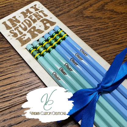 A set of personalized pencils in shades of light blue and mint green, wrapped in a blue ribbon and presented on a wooden card engraved with 'IN MY STUDENT ERA'. The pencils are personalized with the name 'Oliver'.
