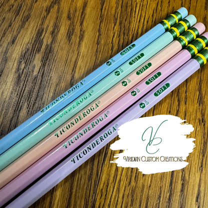 Six Ticonderoga pencils in pastel blue, mint, peach, pink, lavender, and light purple are arranged diagonally on a wooden surface. A white logo overlay for 'Viridian Custom Creations' is in the bottom right corner.